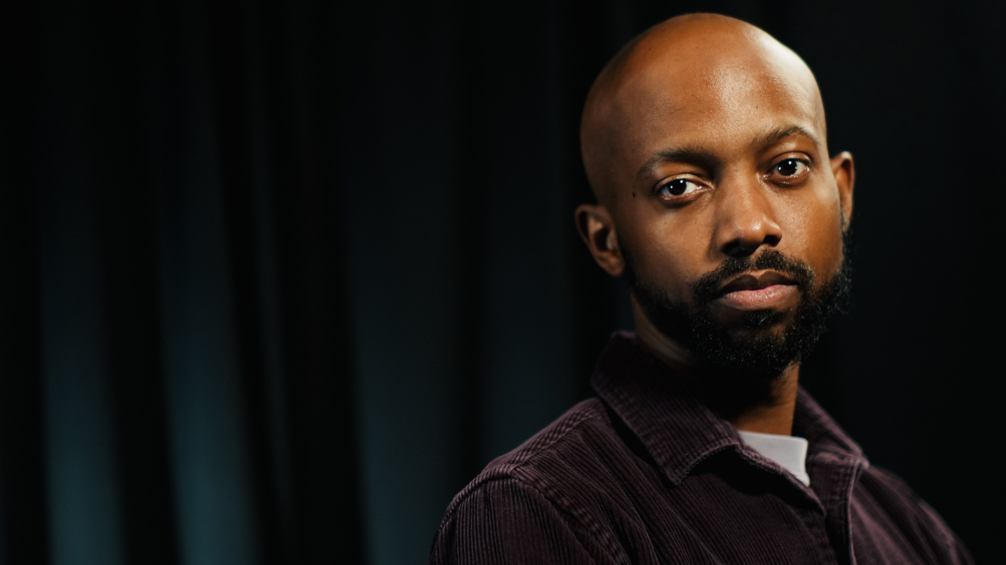 Portrait of Davone Bonneau Jr. against a dark curtain, looking into the lens