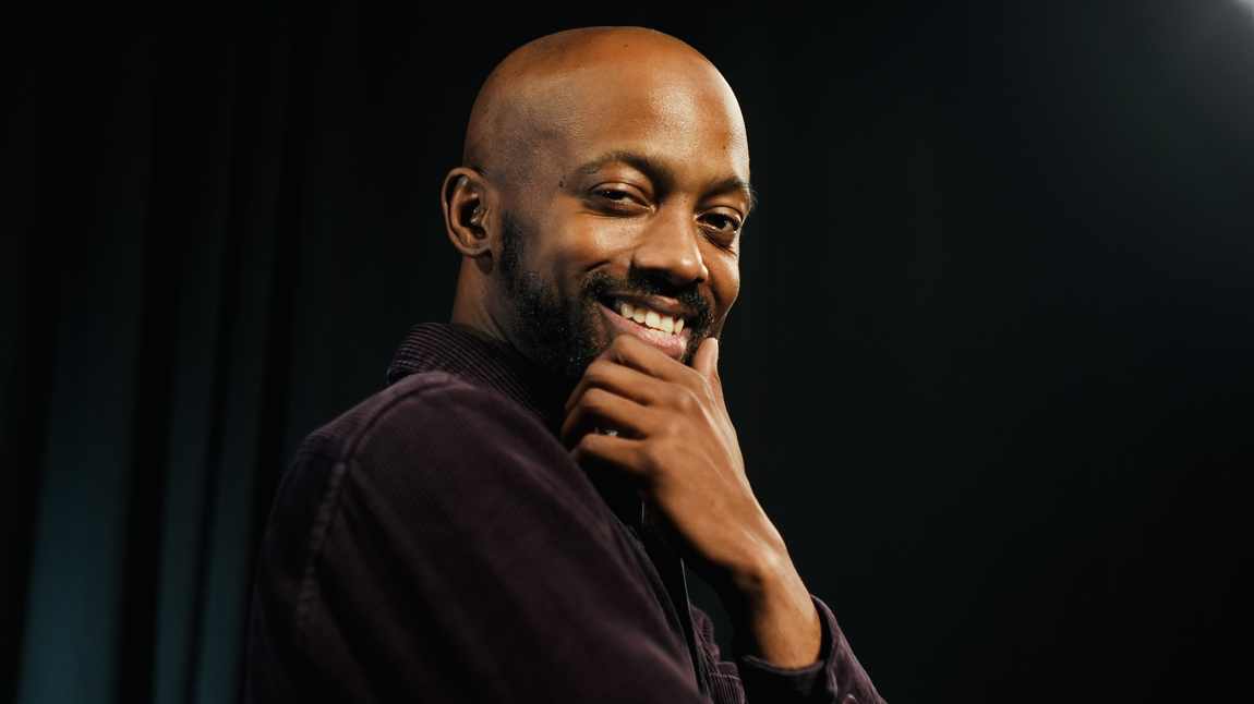 Davone smiling, hand on chin, against a dark curtain